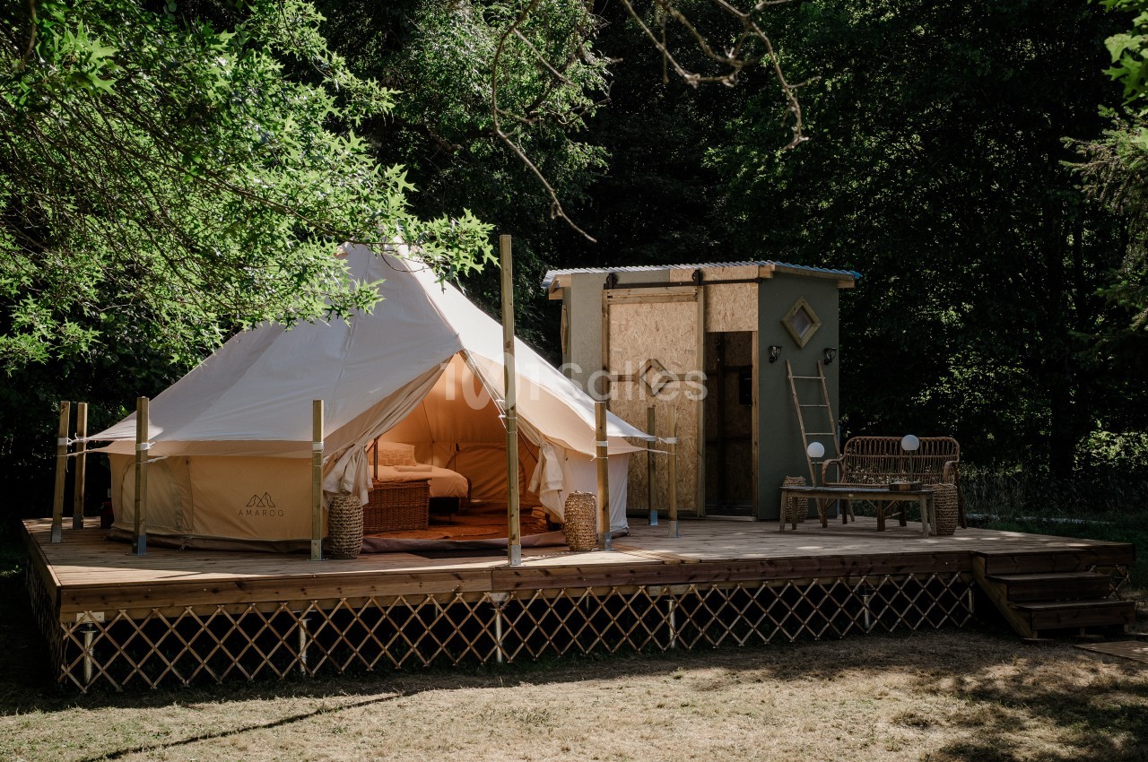 Tente de type bell tent en toile claire installée sur une terrasse en bois surélevée, à l'ombre d'arbres, avec à côté un petit bloc sanitaire en bois et une banquette en rotin. Wait, I must write in French only and check length: "Tente de type bell tent"