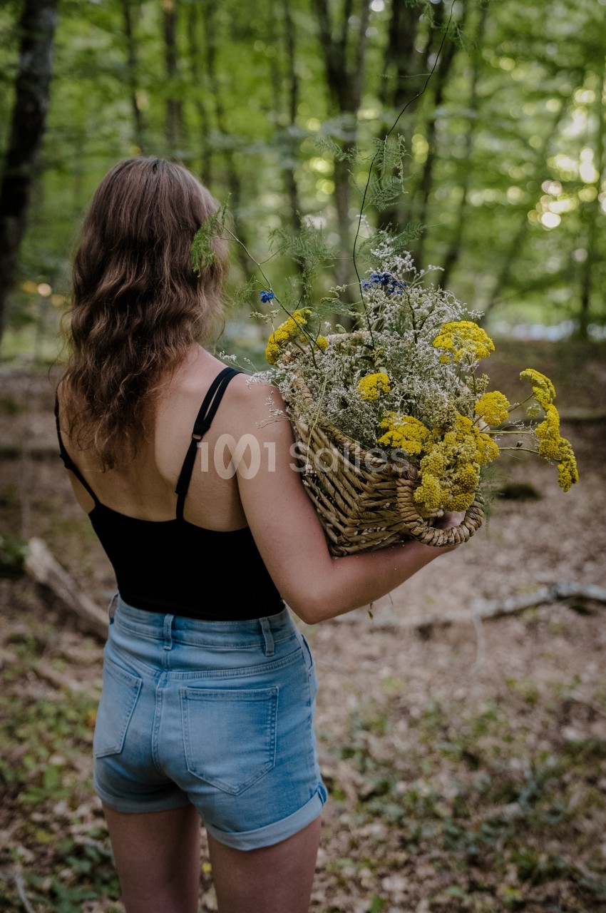Femme de dos en tenue légère, tenant un panier en osier rempli de fleurs jaunes et blanches dans une forêt verdoyante.