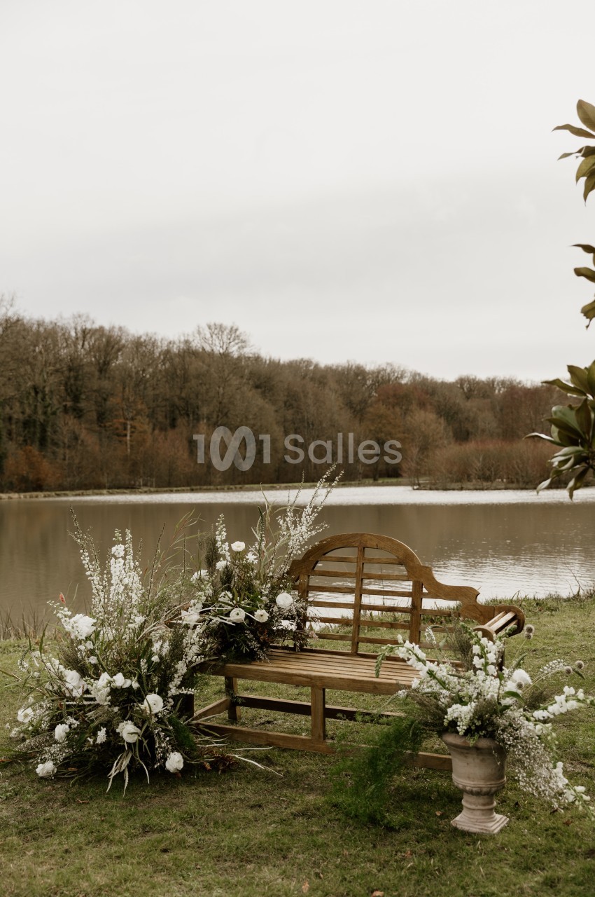 Banc en bois orné de fleurs blanches et de verdure, disposé sur l'herbe au bord d'un étang, devant une lisière d'arbres dénudés.