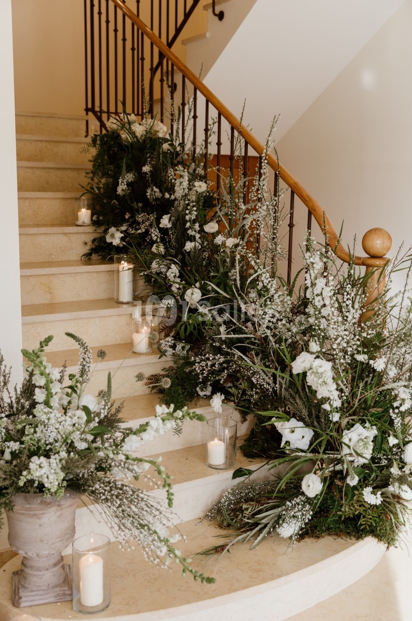 Escalier en pierre clair bordé d'une rampe en bois, orné de compositions florales blanches et vertes et de bougies en verre disposées sur les marches.