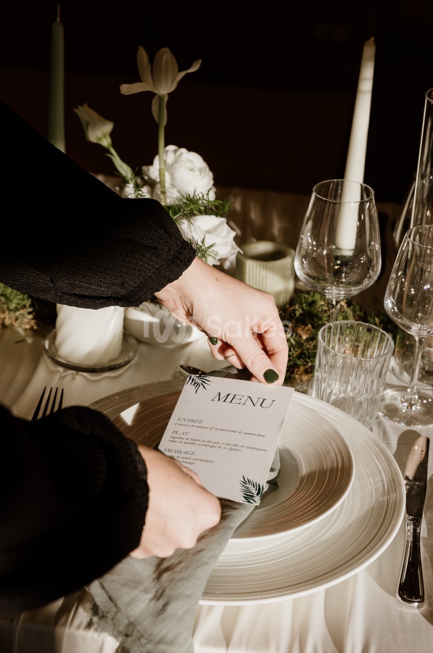 Table dressée avec une carte de menu posée sur une assiette, entourée de bougies blanches, de fleurs et de verres.
