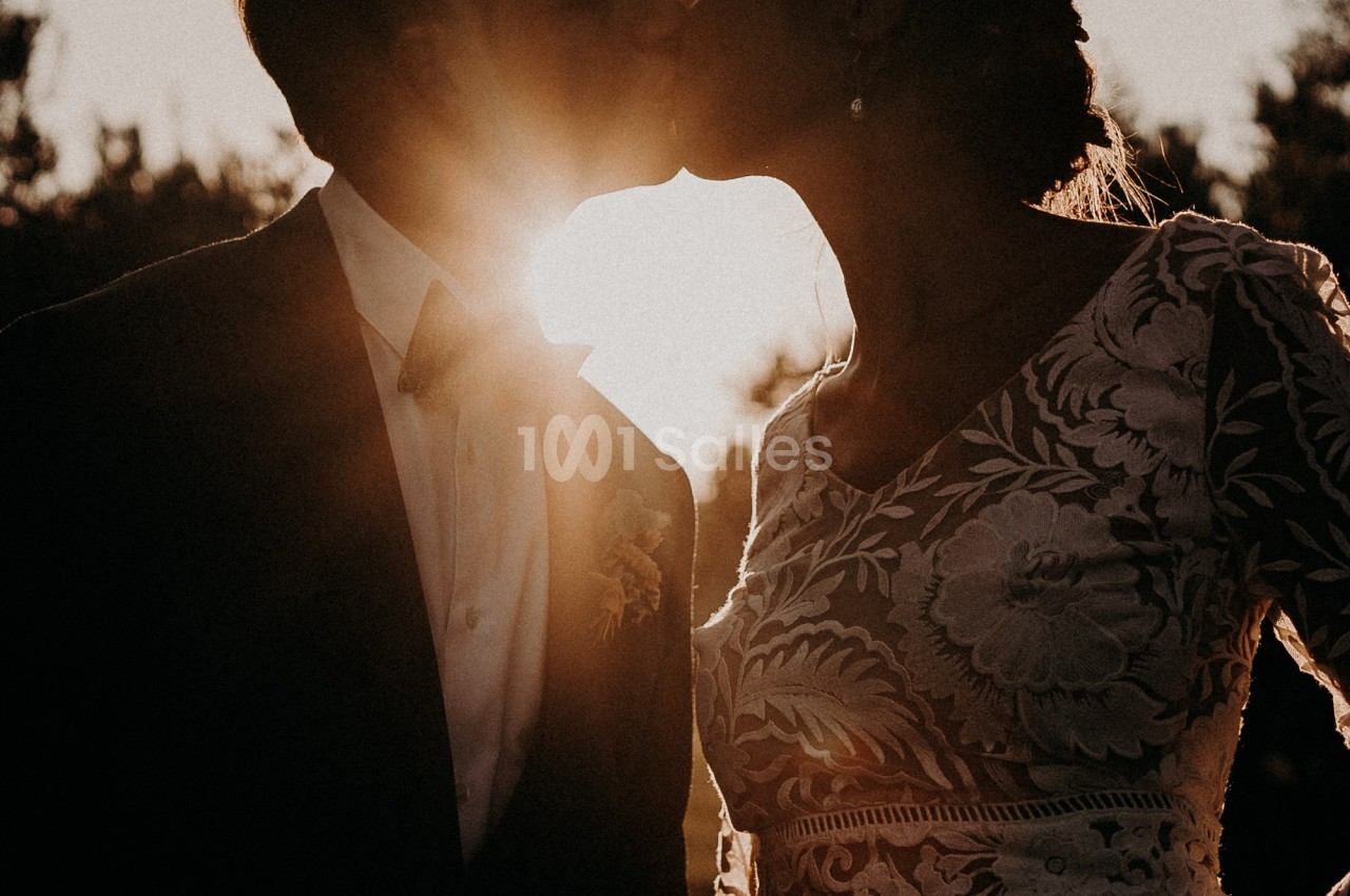Un couple de mariés de dos, enlacés face à un soleil couchant qui éclaire à contre-jour; le marié porte un costume sombre et la mariée une robe en dentelle fleurie. Wait, I must write in French only and the output must be one sentence, under 125