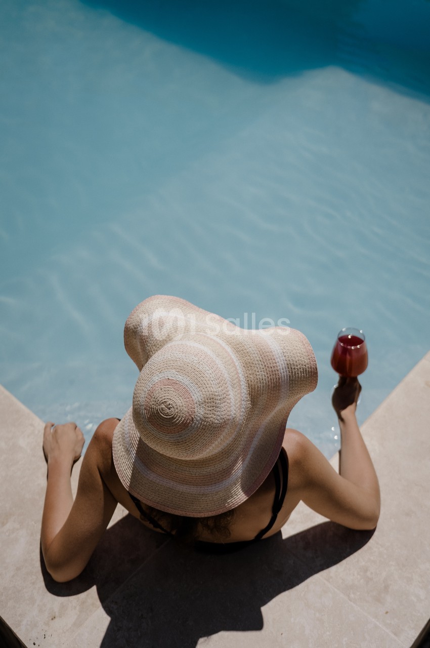 Personne vue de dos, chapeau de paille à larges bords, assise au bord d'une piscine et tenant un verre de vin rosé, sous un fort ensoleillement.
