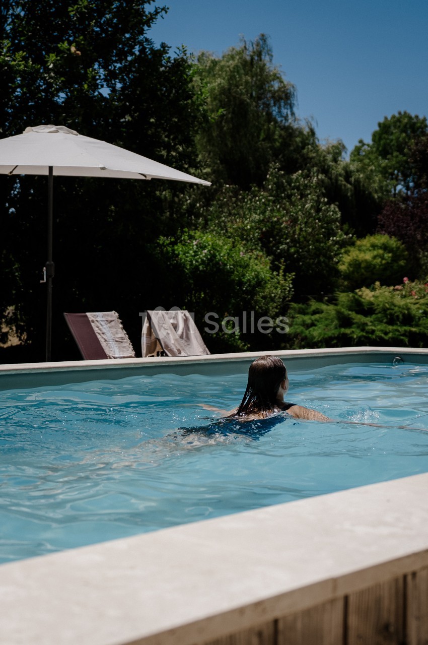 Une personne nage dans une piscine extérieure rectangulaire bordée de pierre claire, avec un parasol blanc et des transats à l'ombre d'arbres.
