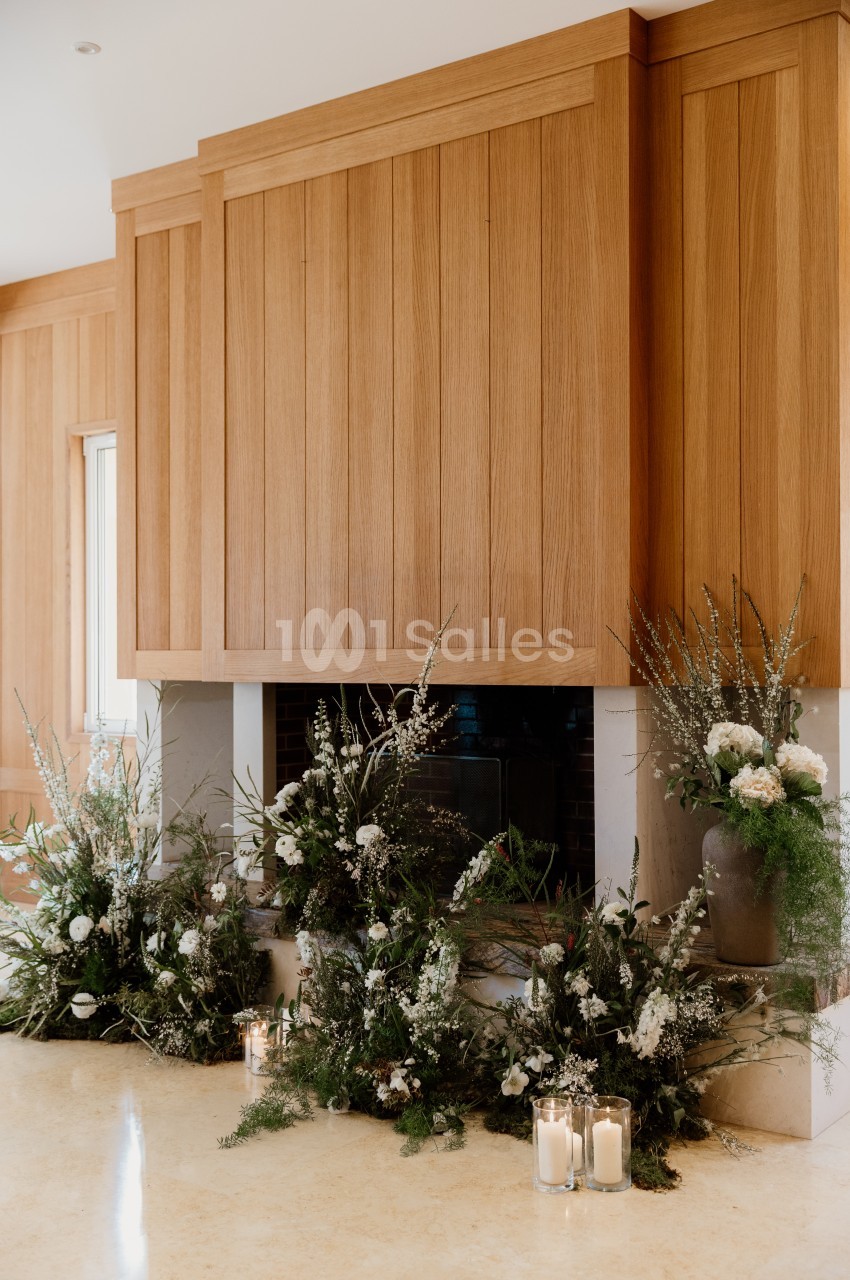 Cheminée en bois clair à panneaux verticaux, ornée au sol de grands bouquets blancs et verts et de bougies en verre, dans une pièce au sol clair.