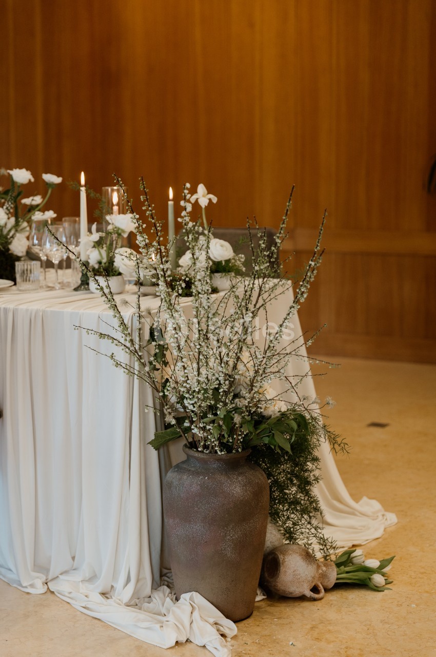 Table nappée de blanc dressée de bougies, verres et fleurs blanches, avec un grand vase en grès rempli de branches fleuries devant un mur en bois.