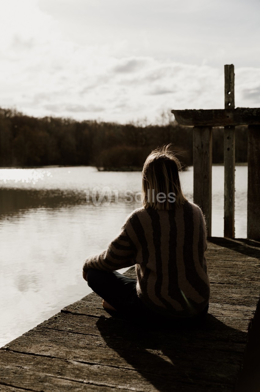 Une jeune femme de dos, assise en tailleur sur un ponton en bois, face à un lac au bord duquel se dressent des arbres, sous un ciel nuageux.