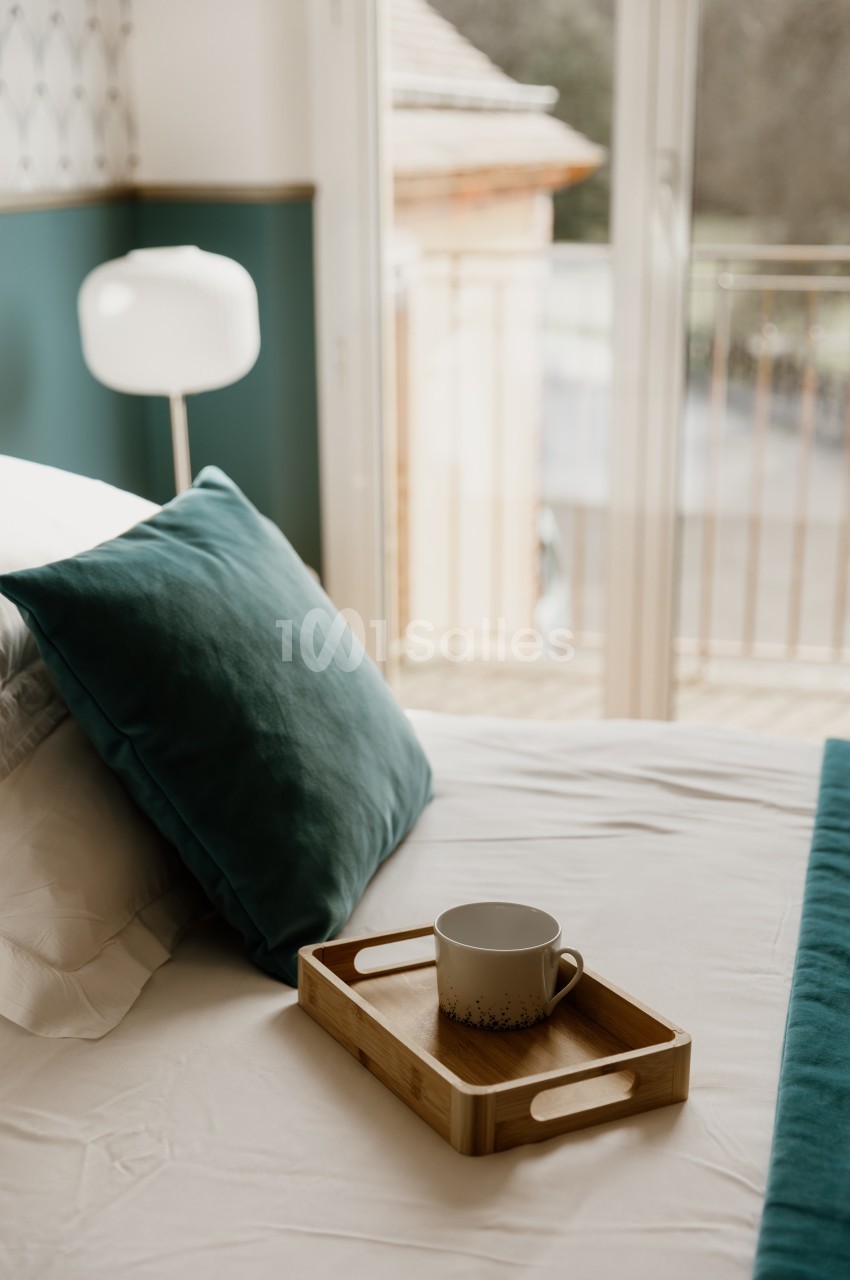 Chambre au lit recouvert de draps clairs, avec un coussin vert sapin et un plateau en bois portant une tasse, près d'une porte-fenêtre ouverte sur un balcon.
