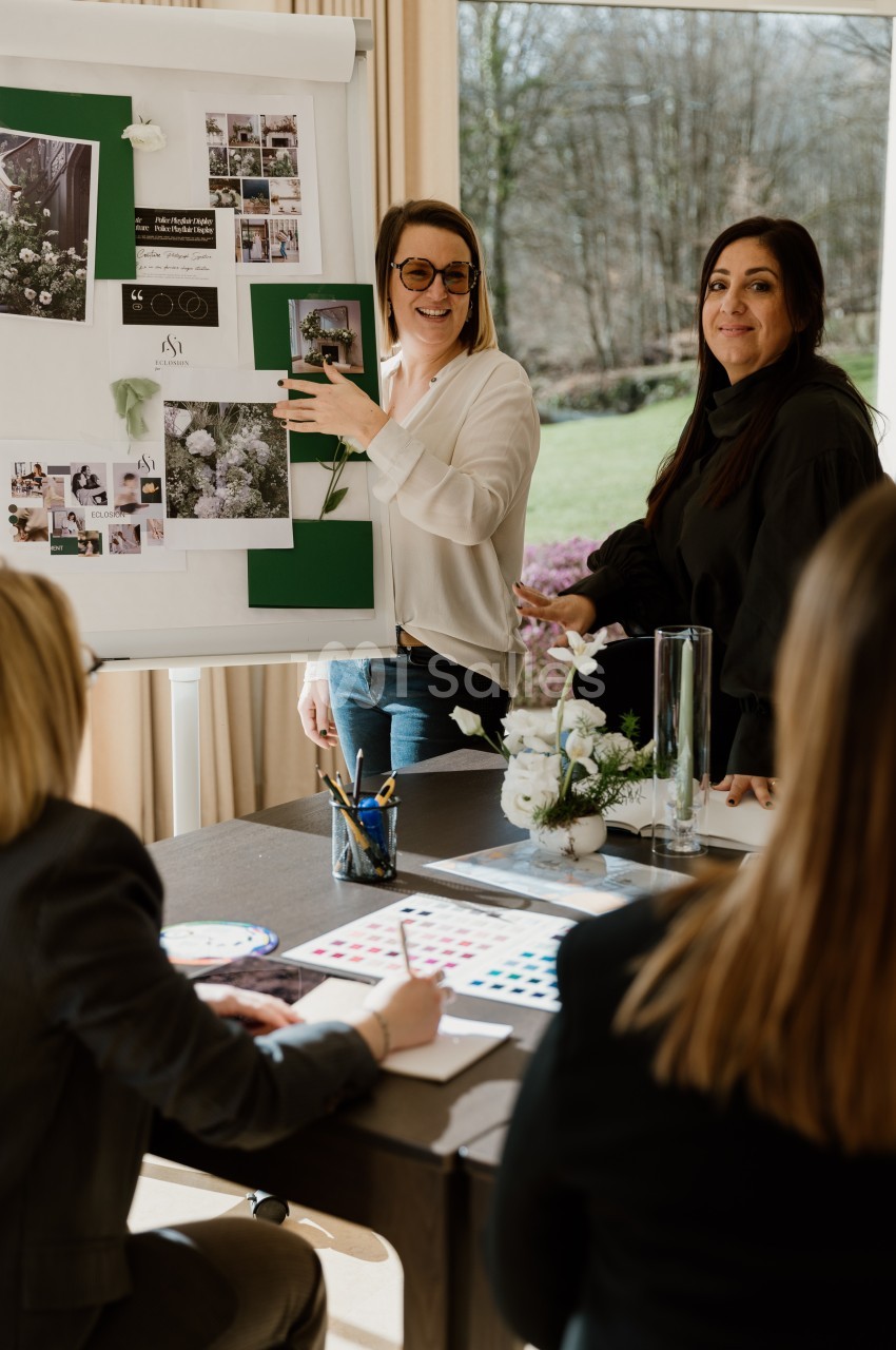Deux femmes présentent un tableau d'inspiration vert et blanc à une table, avec un bouquet de fleurs blanches et des nuanciers posés devant des participants.
