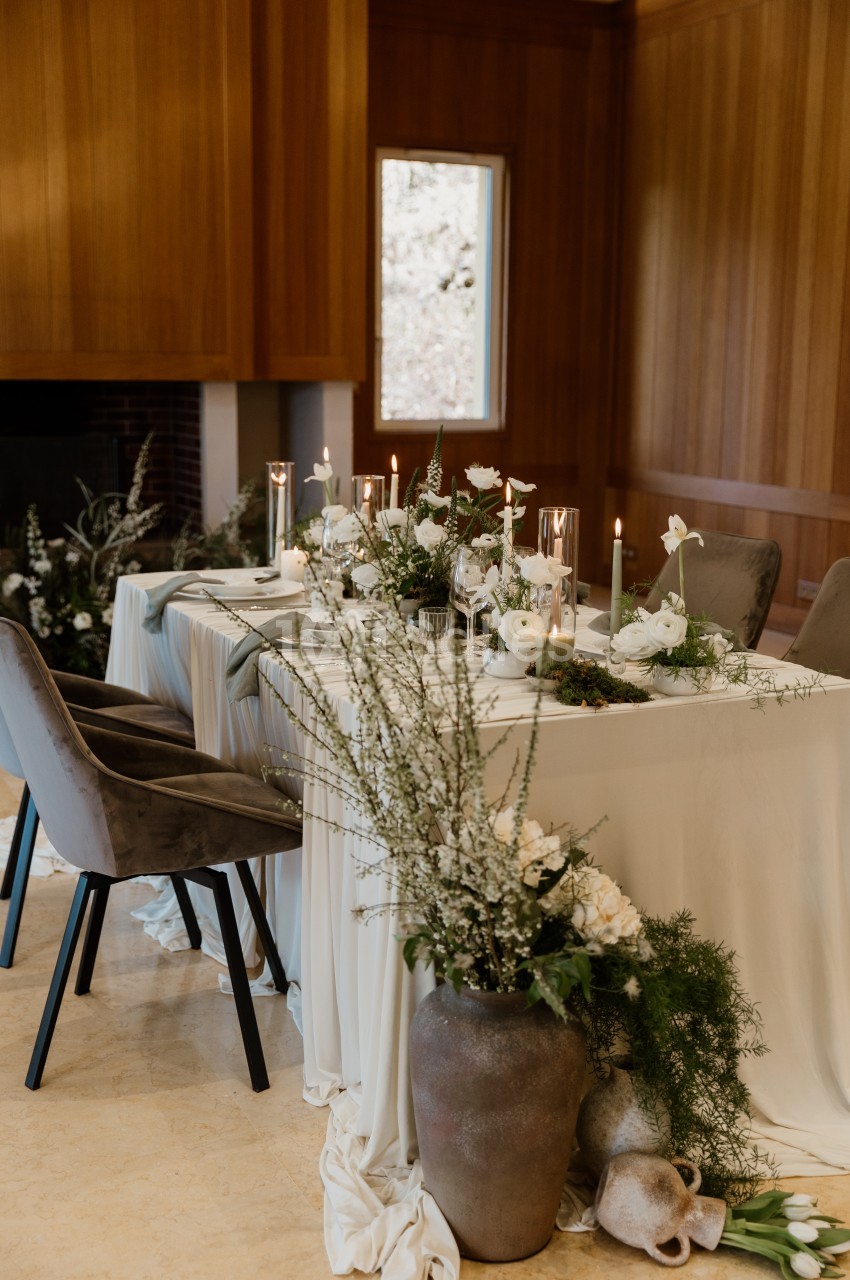 Table dressée en longueur, nappe blanche, bougies, fleurs blanches et vase en grès au premier plan, dans une salle aux murs boisés.