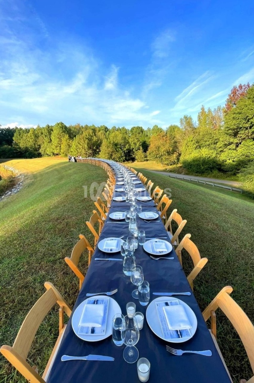 Table longue dressée en extérieur sur une pelouse en pente, nappe bleu marine, assiettes blanches, verres et bougies, bordée de chaises en bois sur plusieurs dizaines de couverts. Let me check constraints: one sentence, under 125 chars? Count: "Table