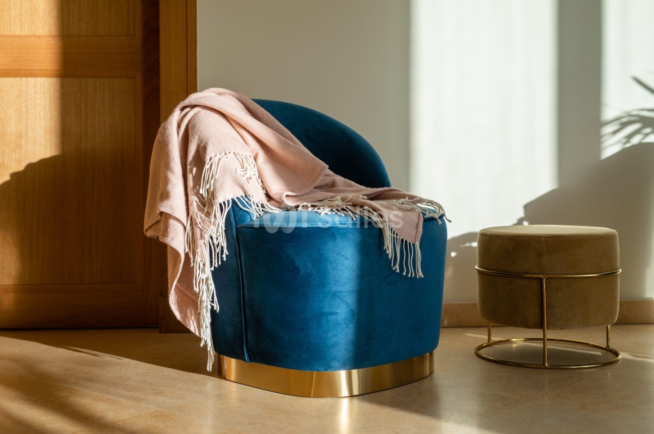 Fauteuil arrondi en velours bleu canard à base dorée, recouvert d'un plaid rose pâle à franges, dans une pièce éclairée par la lumière du jour, à côté d'un pouf doré. Let me verify length: roughly 170 characters, too long. Need ≤125. Fauteuil rond en