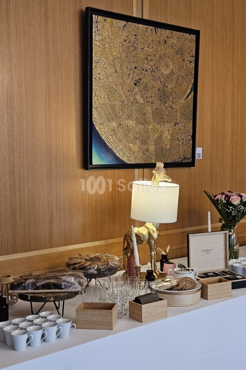 Buffet dressé avec tasses blanches, verres, pâtisseries sous cloche, fleurs et lampe dorée devant un grand tableau doré sur panneau de bois clair.