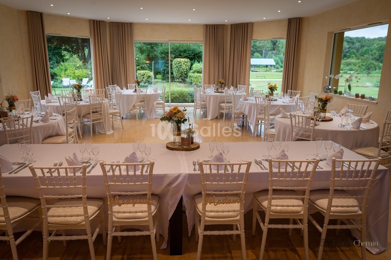 Salle de réception avec tables rondes et table longue nappées de blanc, ouverte sur un jardin par de grandes baies.
