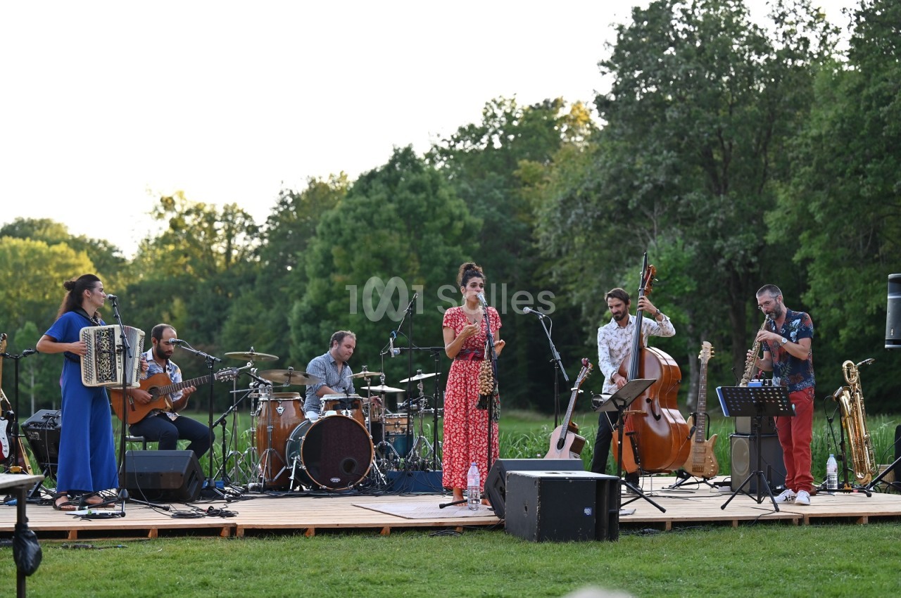 Groupe de musique en plein air sur une scène en bois, avec accordéon, guitare, batterie, contrebasse, saxophones et chanteuse, entouré d'arbres.