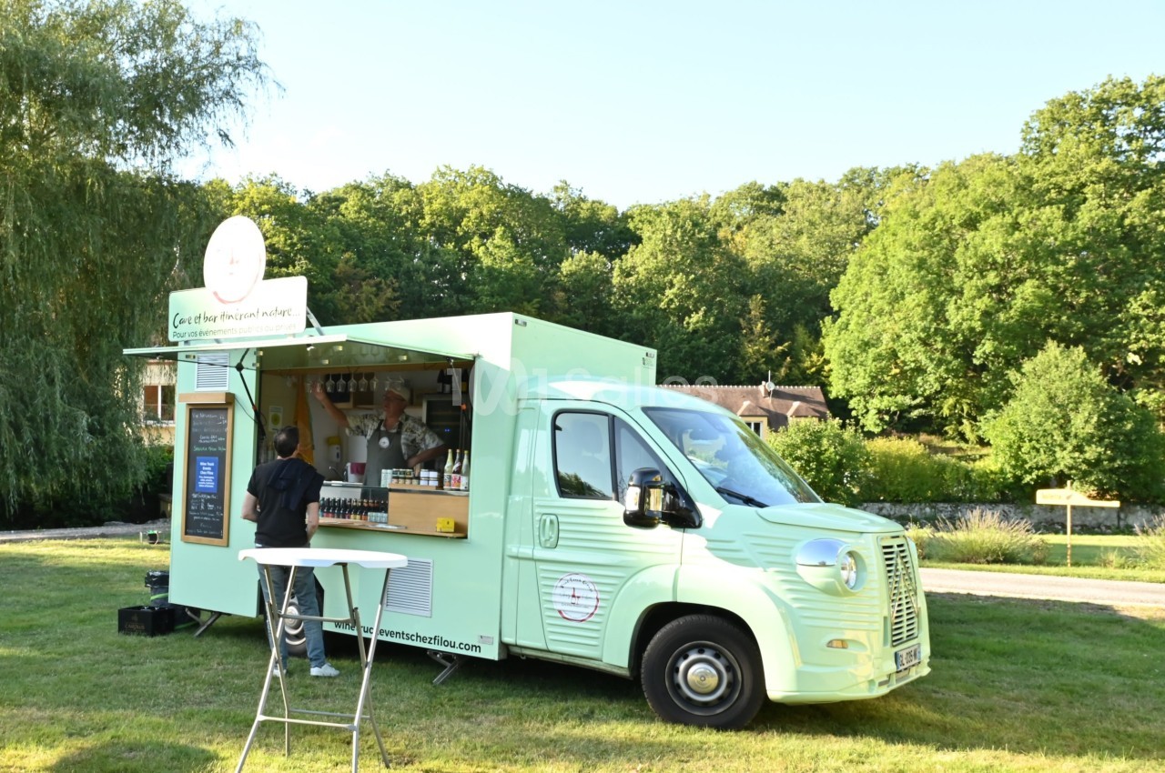 Food truck vert menthe garé sur une pelouse, avec un serveur derrière un comptoir ouvert et un client debout devant, sous de grands arbres. Note: the character count is about 130; let me shorten. Food truck vert menthe garé sur une pelouse, un barman