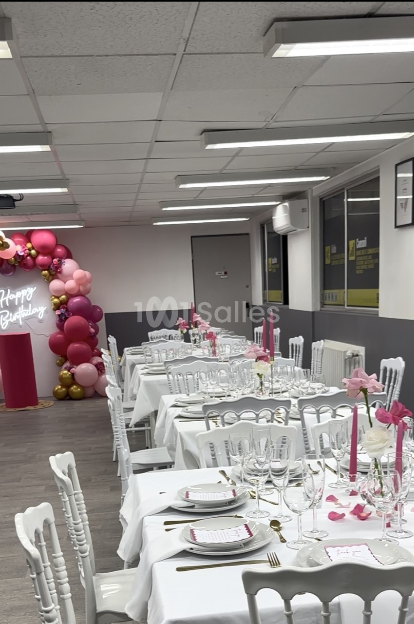 Salle aménagée pour réception avec tables dressées, chaises blanches et décoration rose-burgundy - arche de ballons et guirlande lumineuse.