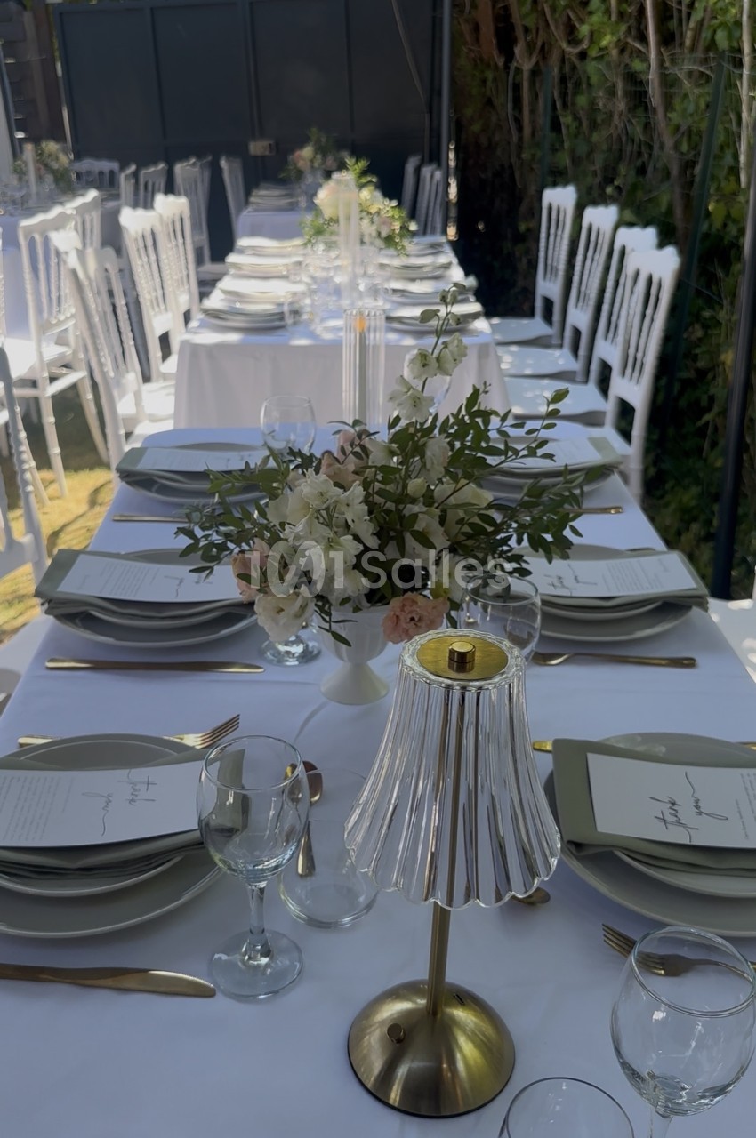 Table dressée en longueur avec nappe blanche, chaises blanches alignées, assiettes empilées, verres, couverts dorés et compositions florales blanches et vertes.