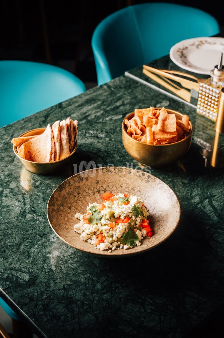Table en marbre vert avec deux chaises turquoise, assiette de couscous garni et bols de chips et crêpes.