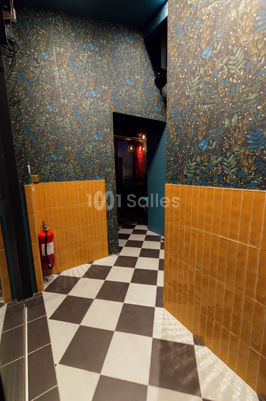Couloir de circulation aux murs tapissés de motifs floraux bleus et or, carrelage damier noir et blanc, boiseries orange sur les parois basses.