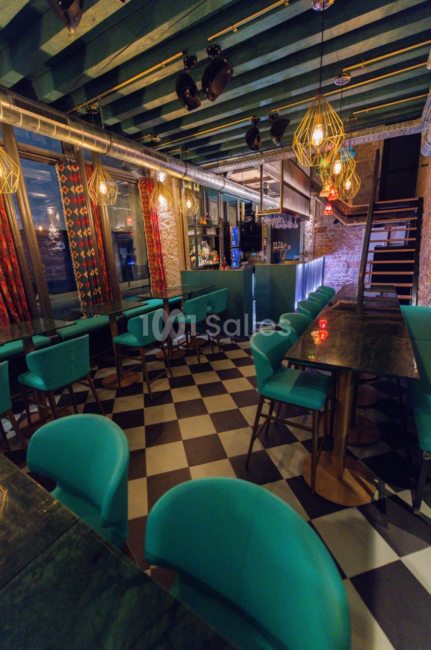 Salle de restaurant au design rétro avec chaises en velours turquoise, sol carrelé noir et beige, bar au fond et luminaires dorés.