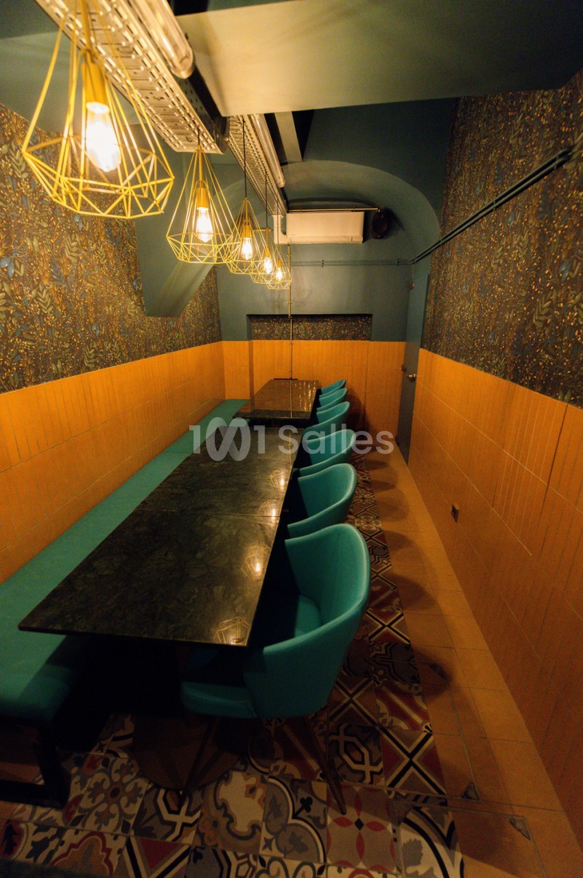 Espace étroit aux murs orange et doré avec table noire et chaises turquoise sous luminaires géométriques.