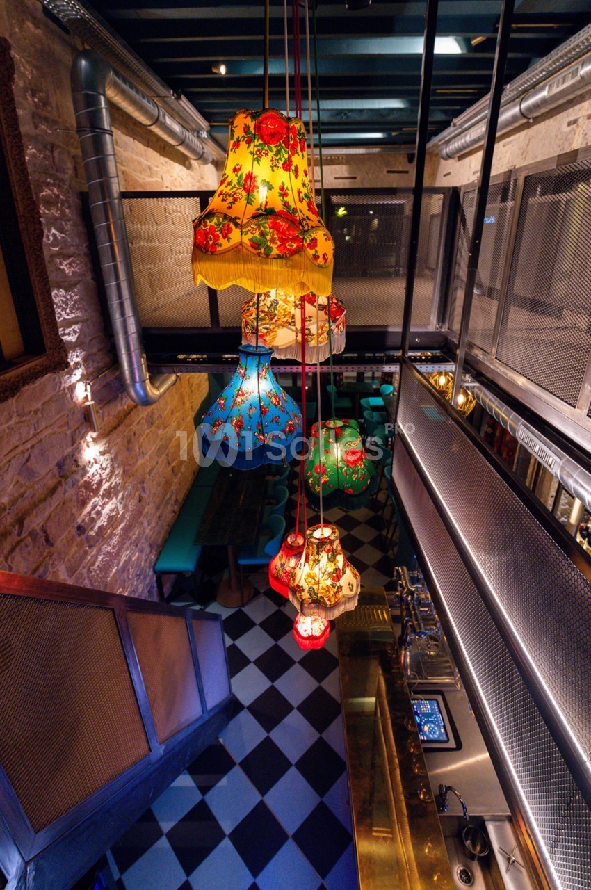 Couloir étroit avec sol à damier noir et blanc, lampes suspendues colorées à motifs floraux, murs en brique et métal.