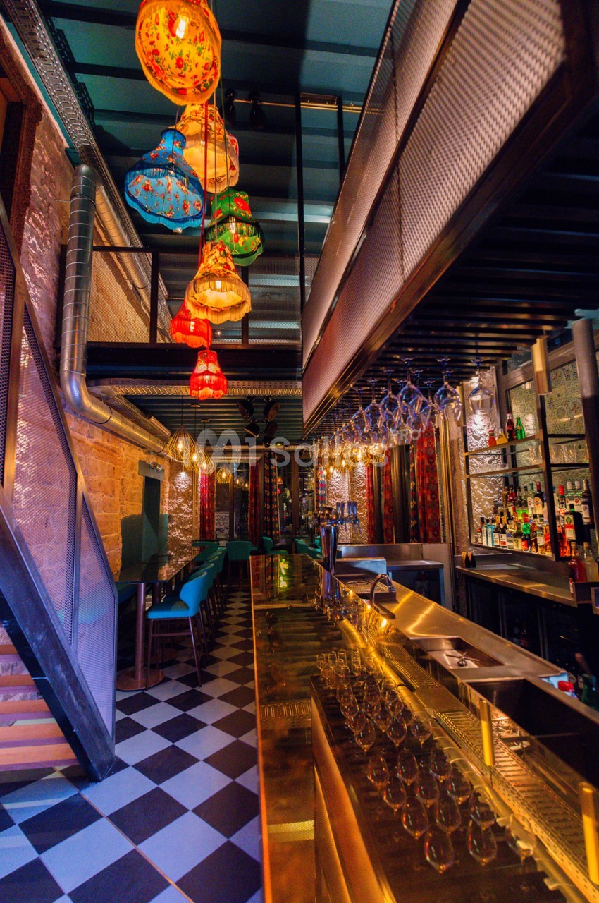 Couloir de bar avec comptoir en inox, sol damier noir et blanc, lanternes colorées suspendues et murs en brique éclairés.