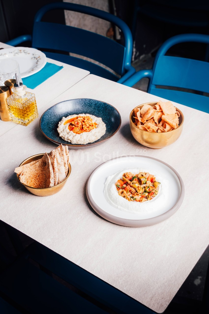 Table dressée avec assiettes de mezze - houmous, pois chiches, pain et crackers, chaises bleues en arrière-plan.