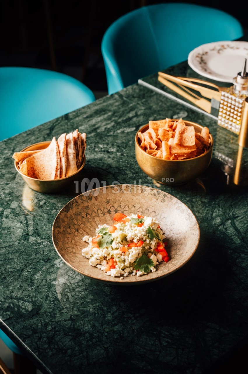 Table en marbre vert avec deux chaises turquoise, assiette de couscous garni et bols de chips et crêpes.
