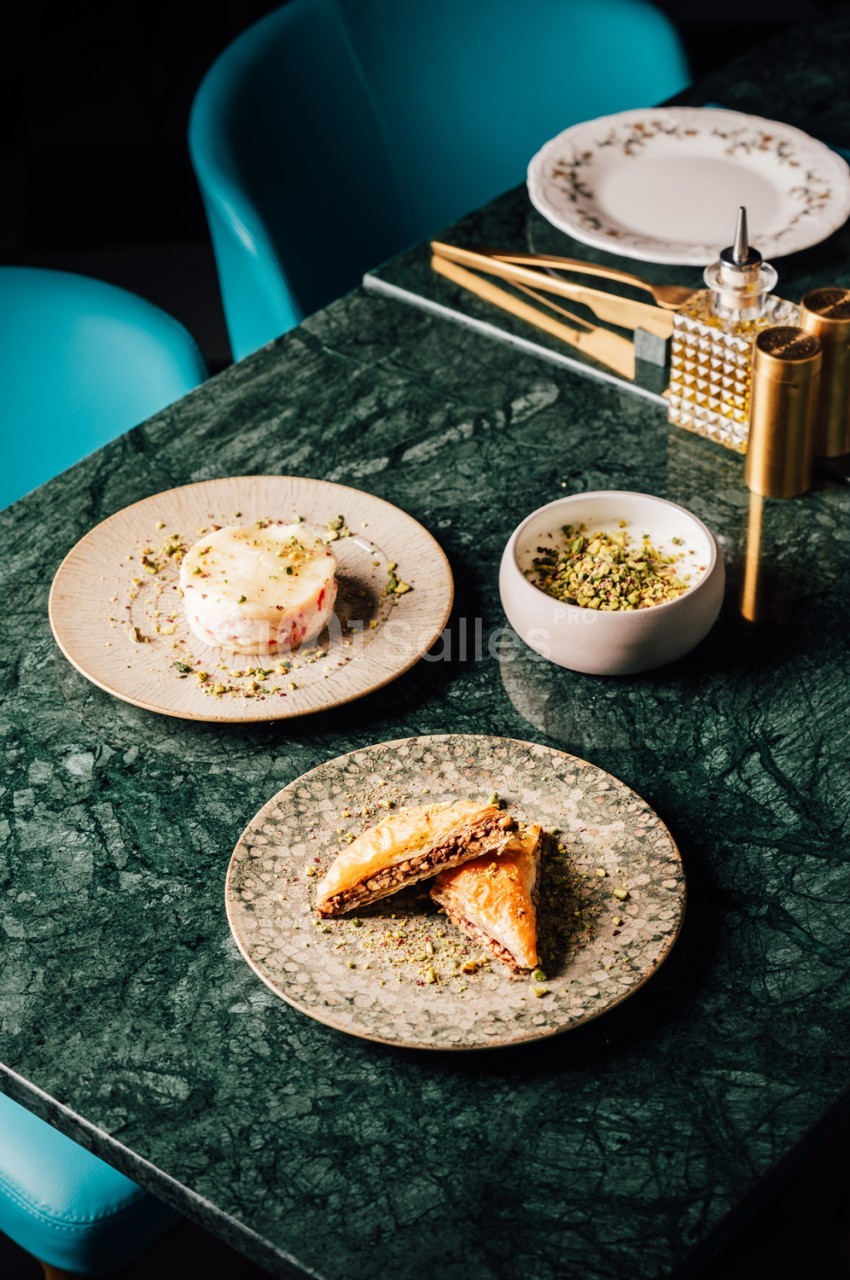 Table en marbre vert avec assiettes de cuisine gastronomique, chaises bleues et accessoires dorés.