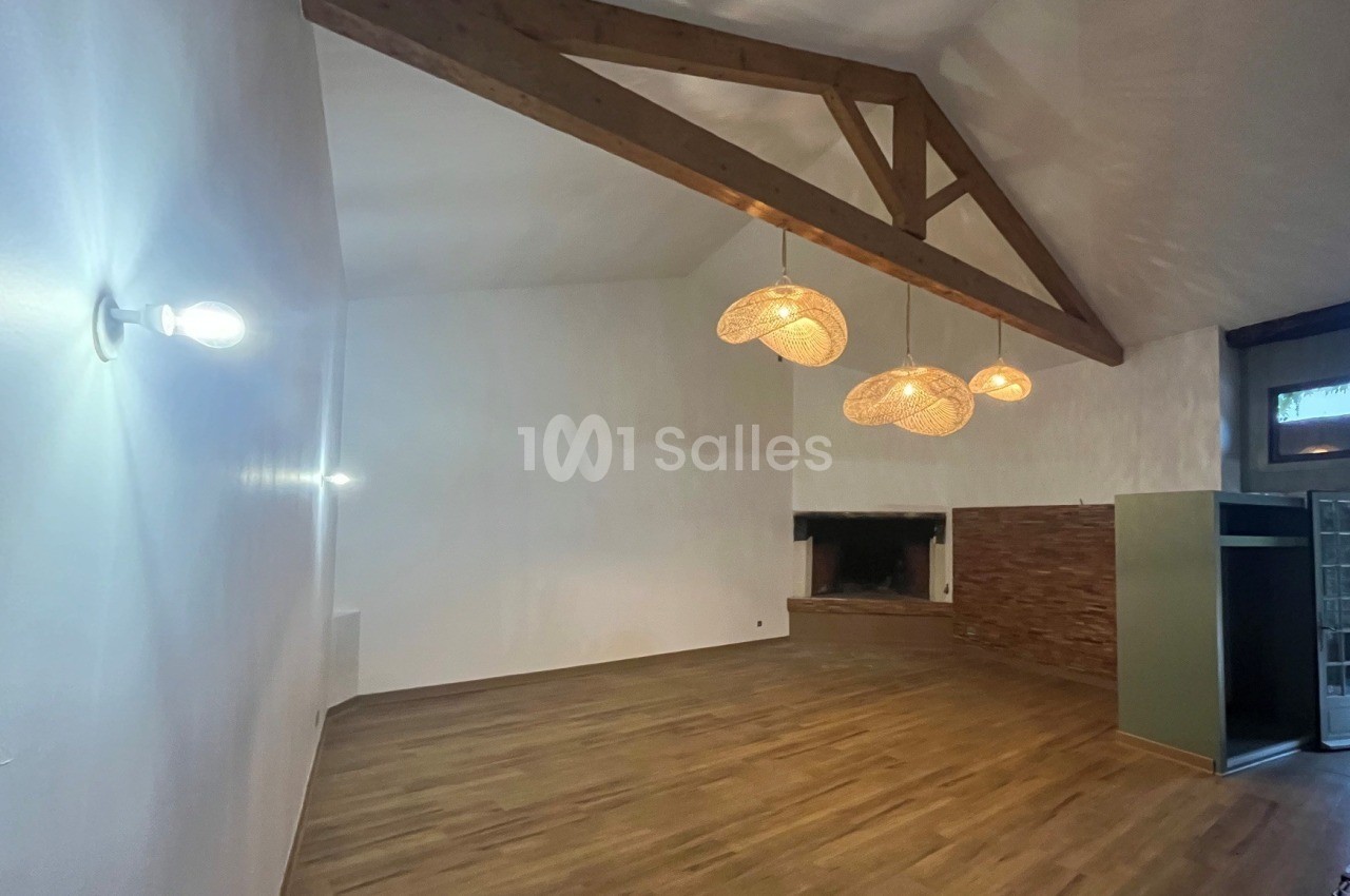 Salle vide au sol en parquet clair, plafond à poutres apparentes en bois, deux suspensions en rotin allumées et une cheminée en pierre à droite.