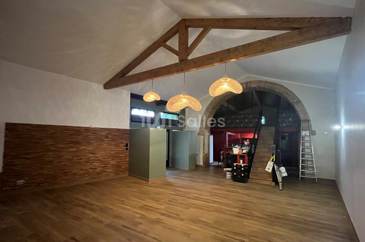 Grande salle vide au sol en parquet clair, plafond à poutres apparentes en bois, suspensions en osier allumées et arche en pierre ouvrant sur un escalier.