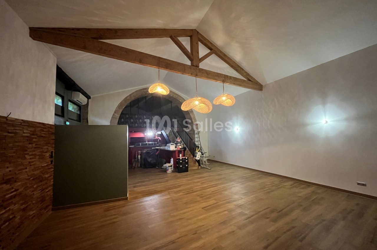 Salle vide au sol en parquet, poutres apparentes, suspensions en osier et arche en pierre donnant sur un espace annexe.