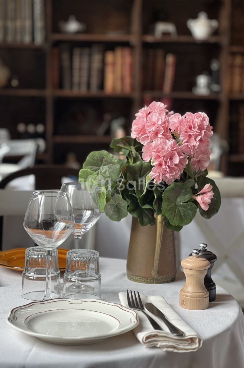 Table dressée avec assiette, verres, couverts, serviette et vase de géraniums roses - bibliothèque en arrière-plan.