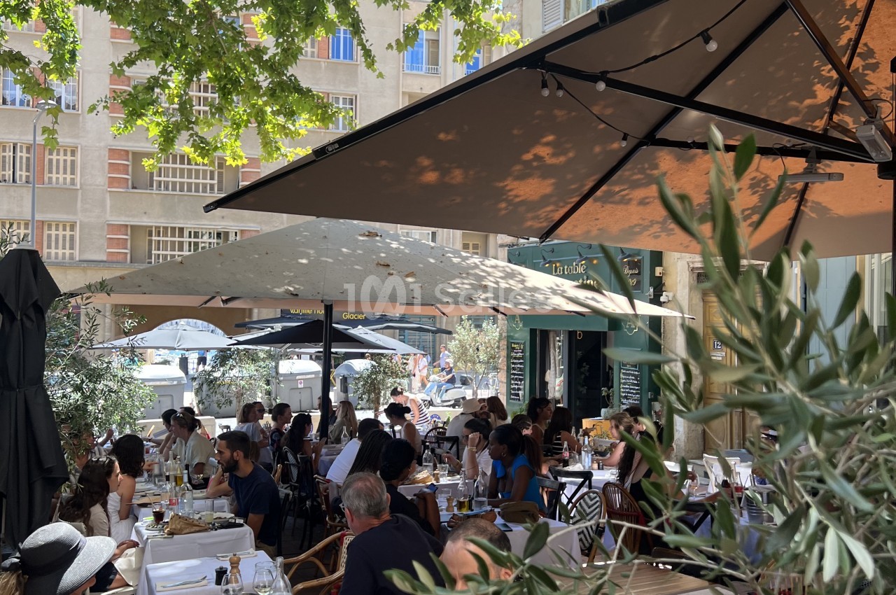 Terrasse ombragée avec tables dressées, convives attablés, façade d'immeuble en arrière-plan.
