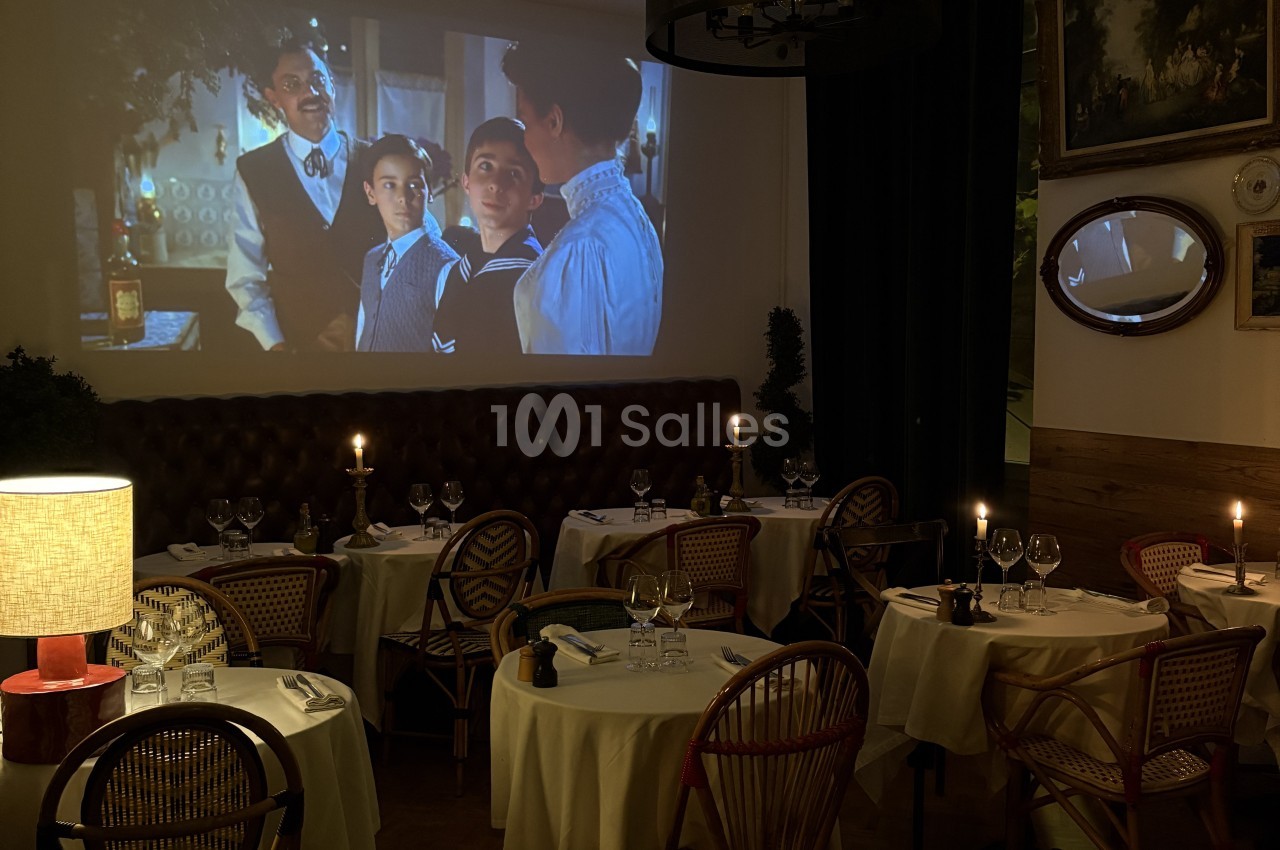 Salle de restaurant avec tables rondes dressées, chaises en rotin, projection cinéma et éclairage tamisé.
