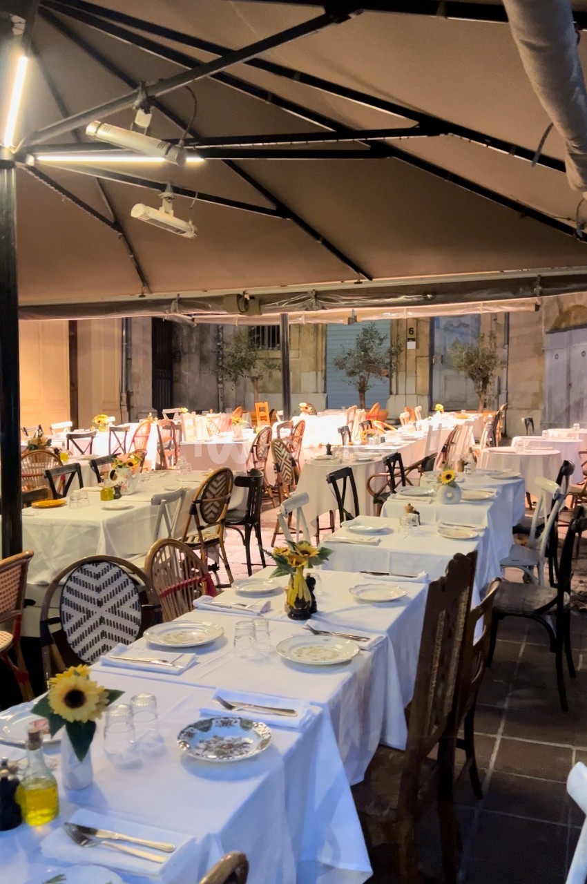 Terrasse couverte aménagée pour un repas avec tables rondes dressées, chaises assorties et décoration florale jaune.
