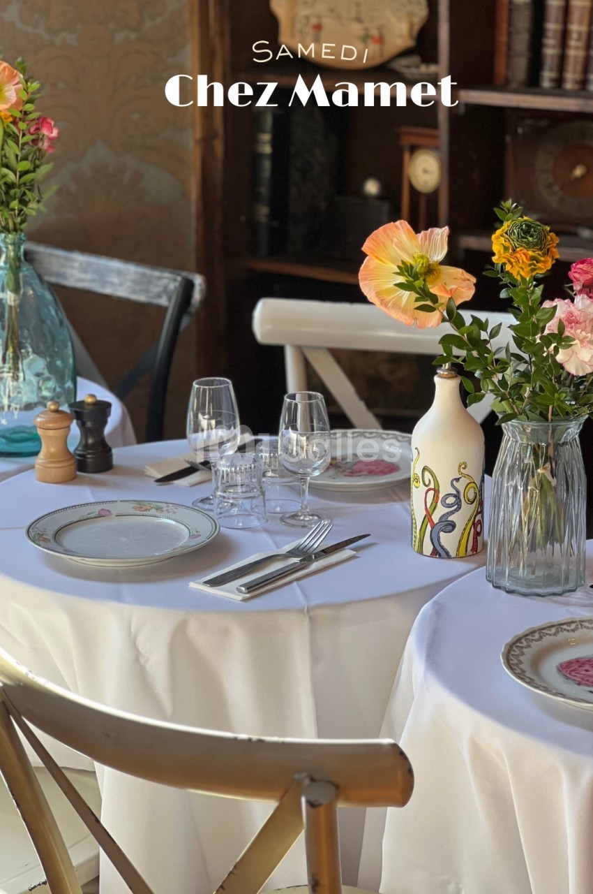 Tables rondes dressées avec nappes blanches, assiettes à motifs floraux, verres et fleurs colorées en vases.