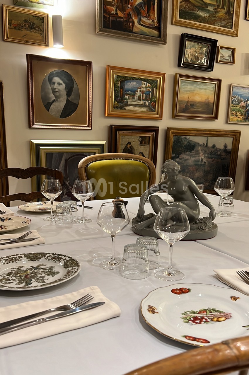 Salle à manger dressée avec table blanche, chaises en bois, verres et couverts - murs ornés de cadres de tableaux anciens.
