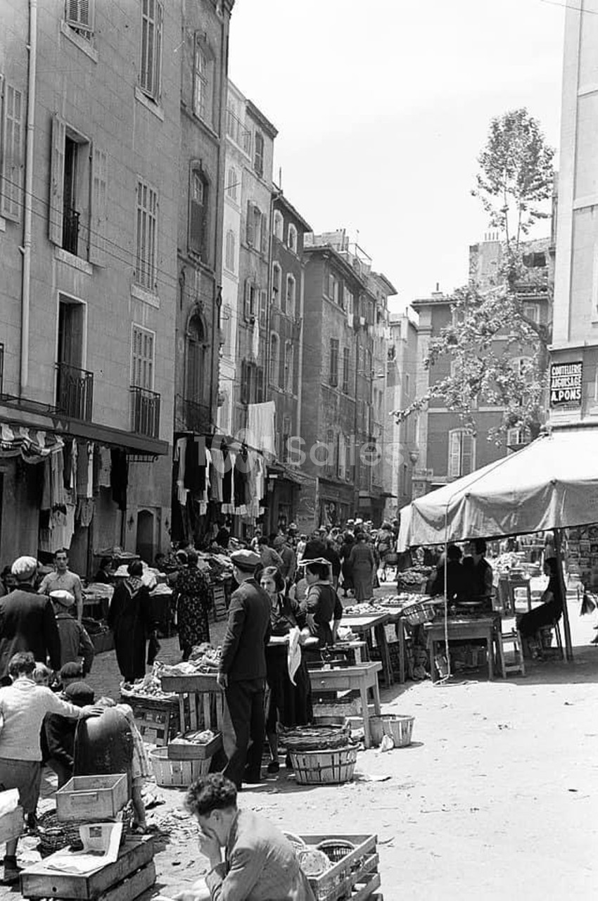 Marché en plein air dans une ruelle de Marseille avec immeubles haussmanniens et étals de fruits et légumes.
