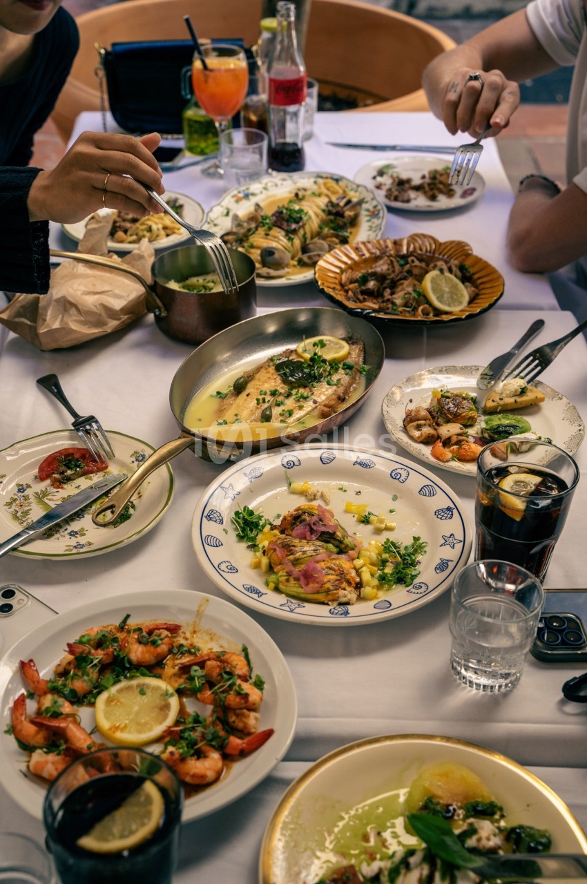 Table dressée avec convives mangeant des fruits de mer et poissons grillés, verres de boissons, paniers et assiettes variées.