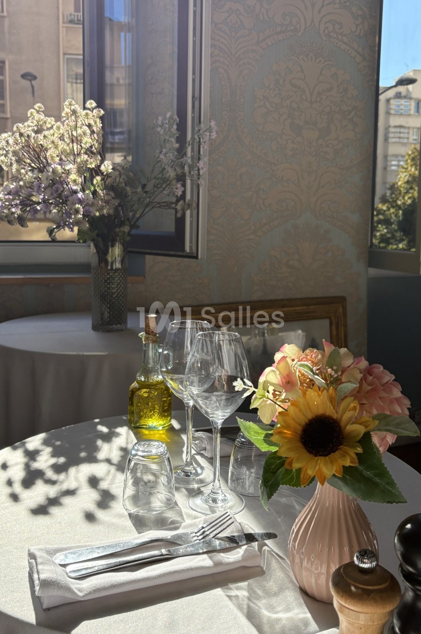Table dressée avec verres, couverts, huile d'olive et fleurs - fond de salle avec baies vitrées donnant sur Marseille.