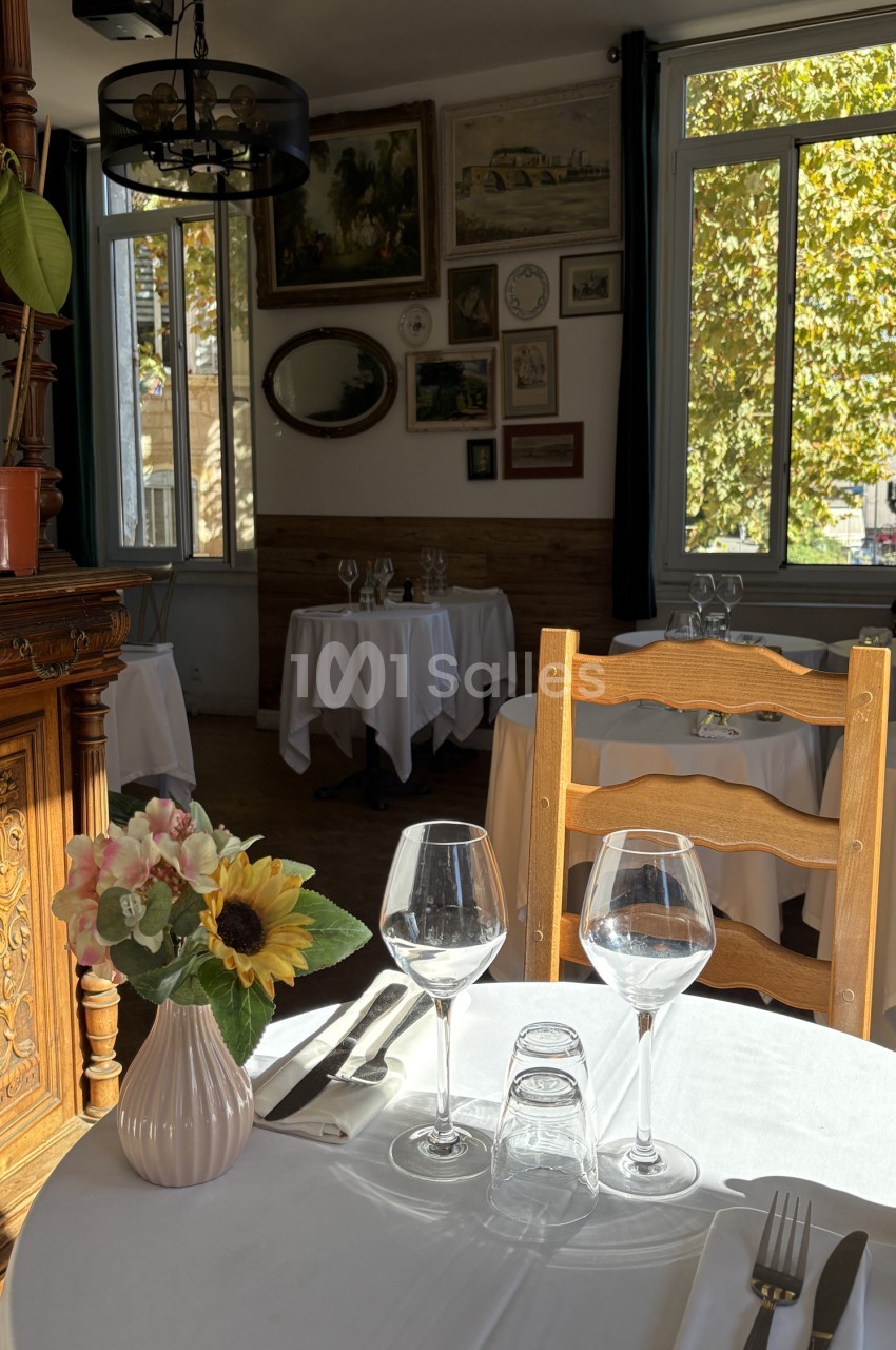 Salle de restaurant avec tables dressées, chaises en bois et murs décorés de cadres, luminosité naturelle des fenêtres.
