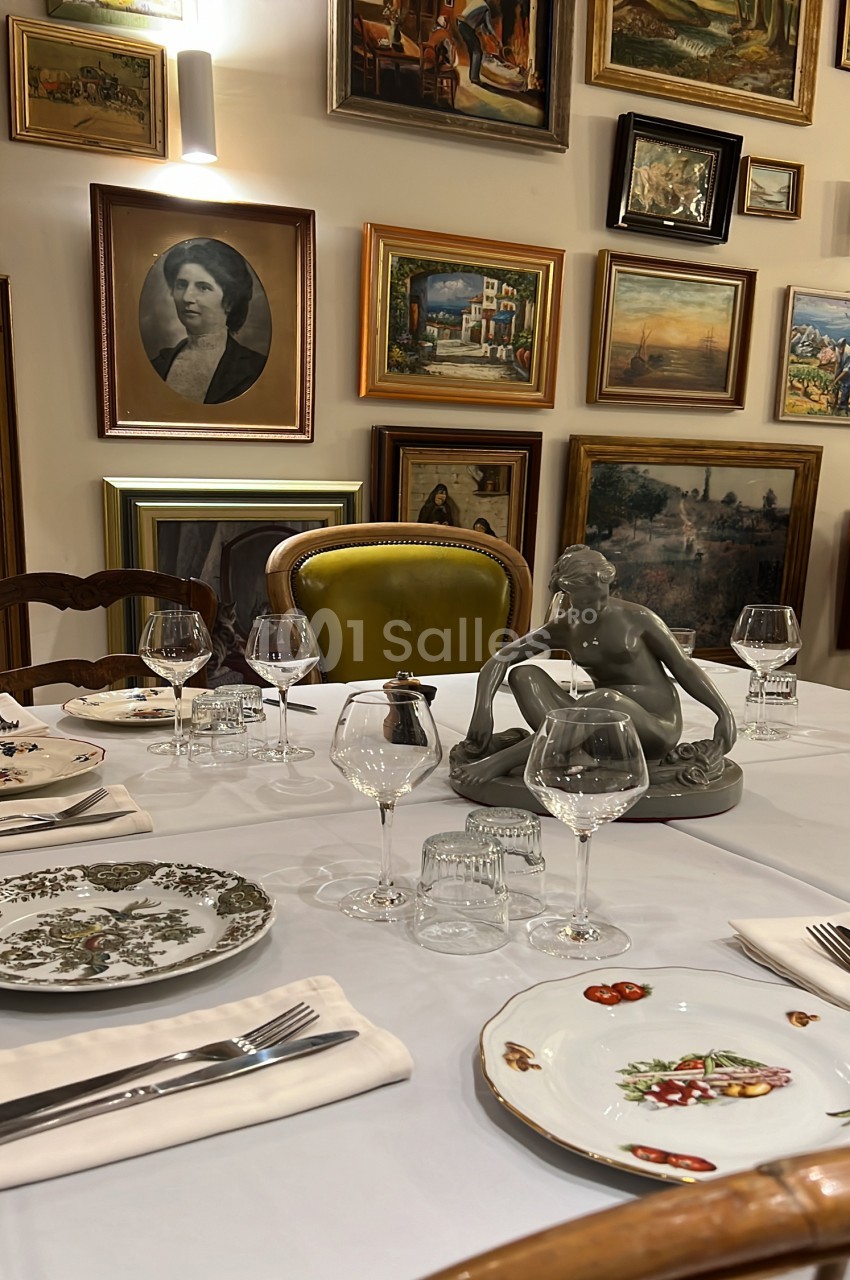 Salle à manger dressée avec table blanche, chaises en bois, verres et couverts - murs ornés de cadres de tableaux anciens.