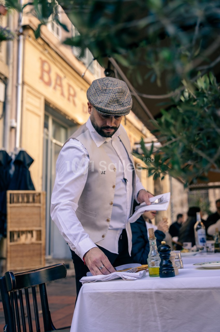 Serveur en gilet clair dressant une table en terrasse devant une façade jaune pâle.