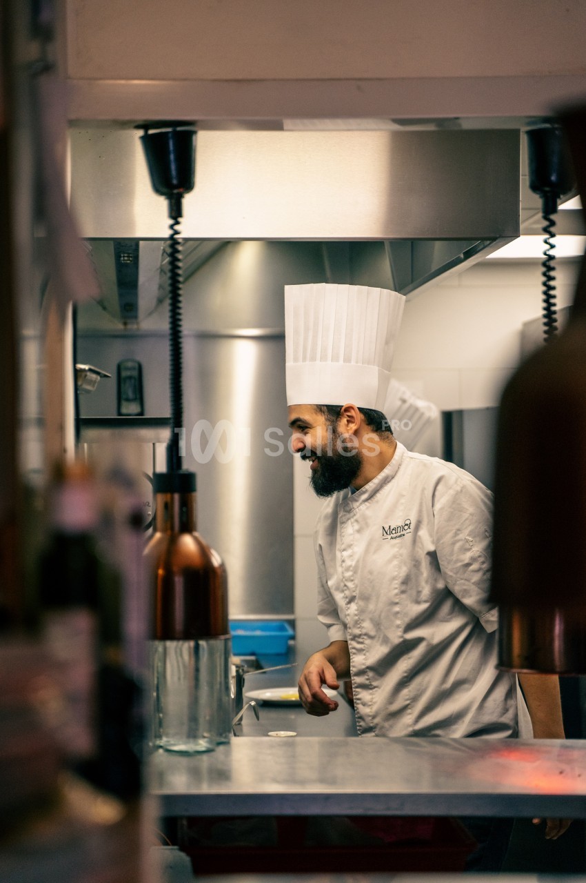 Chef en toque blanche et vêtements professionnels dans une cuisine équipée, lampes chauffantes au-dessus du plan de travail.