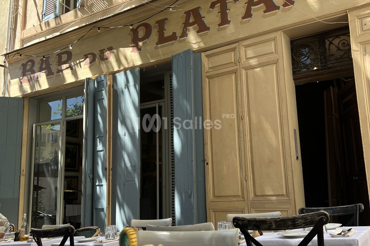Terrasse de restaurant avec tables dressées, chaises noires et façade haussmannienne beige aux portes-fenêtres.