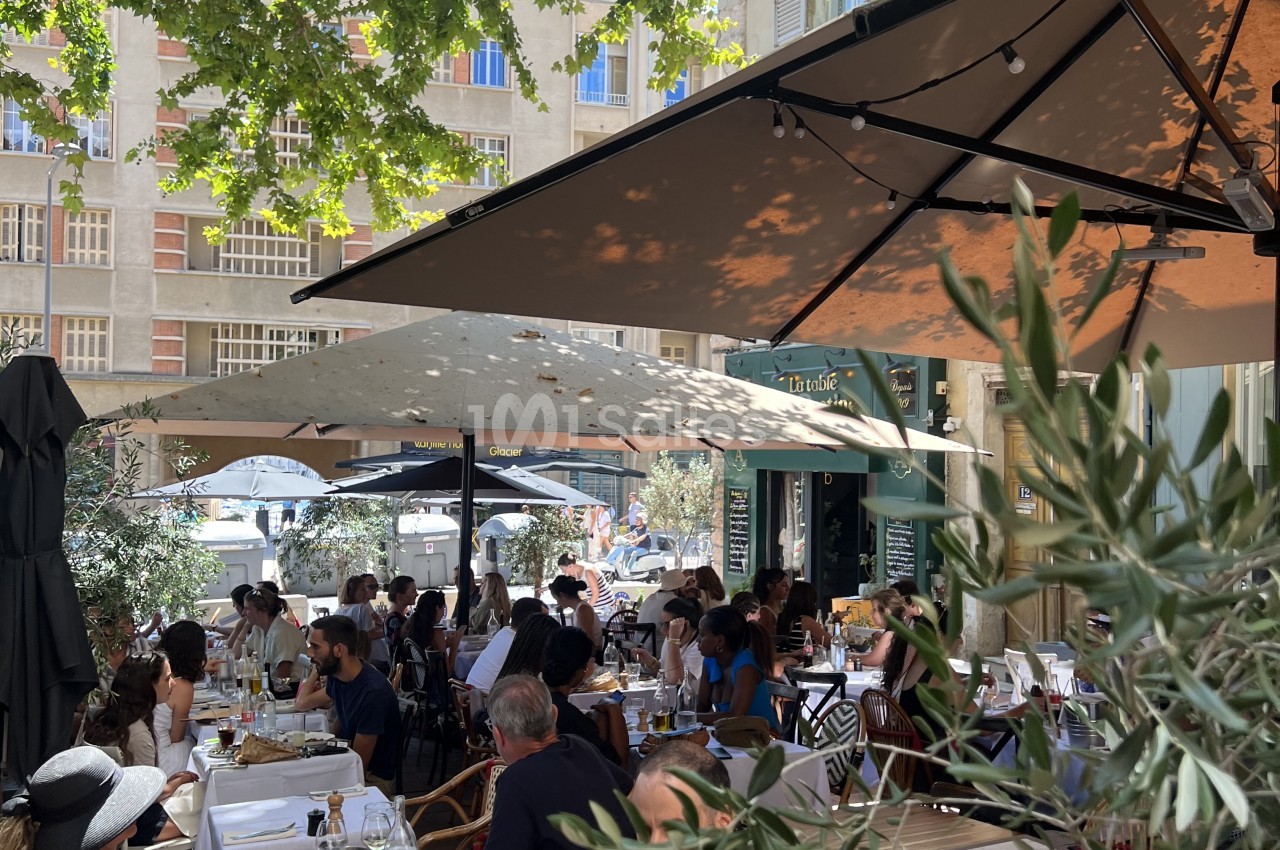Terrasse ombragée avec tables dressées, convives attablés, façade d'immeuble en arrière-plan.