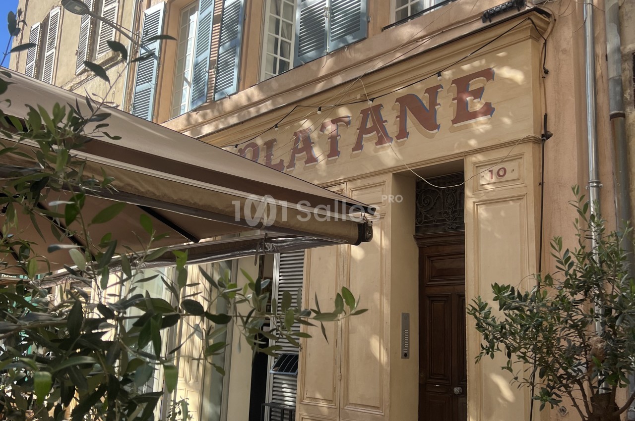 Façade haussmannienne avec volets bleus, auvent rétractable gris et plantes en pot, entrée de restaurant.
